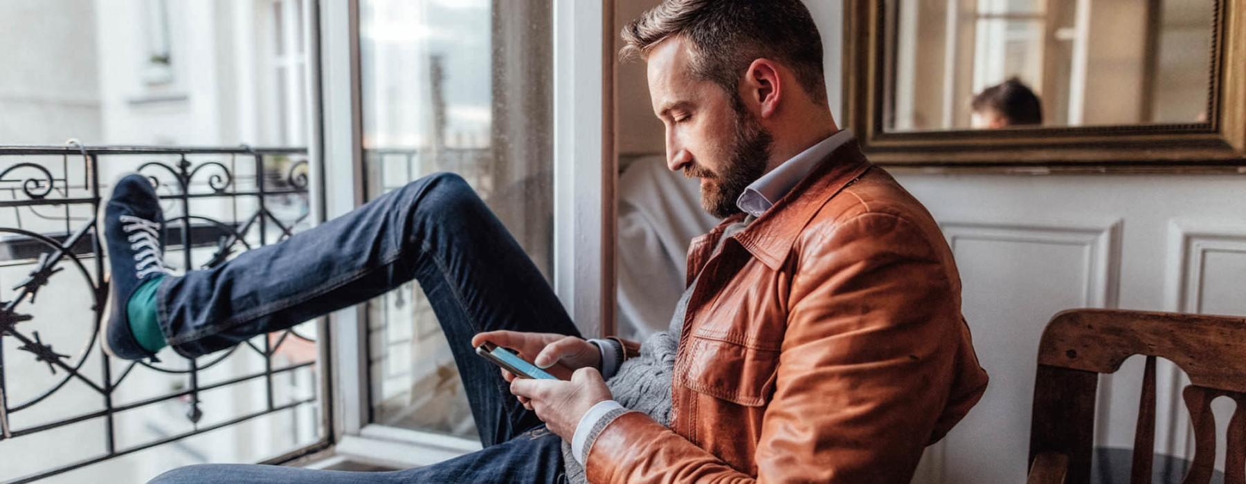 a man sits on a chair and rests his foot on a balcony railing as he looks at his phone in his room
